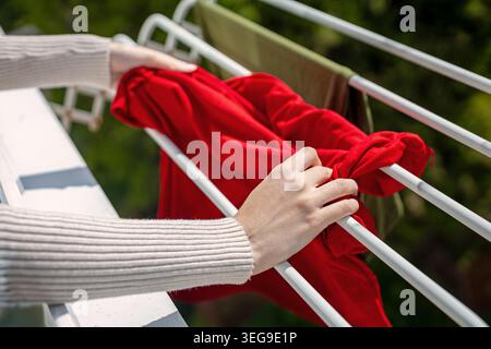 Hanging Red Clothes on Balcony Drying Rack. Hands hanging a bright red garment on an outdoor balcony drying rack in sunny weather. Stock Photo