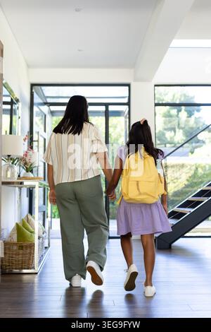 Diverse mother and youth daughter walking in foyer to glass doors holding backpack and water bottle Stock Photo