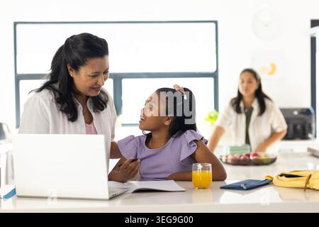 Diverse family leaning and helping youth girl studying at kitchen island with laptop, juice Stock Photo