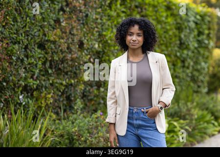 Woman standing beside green hedge on path, wearing beige blazer, jeans, watch, copy space Stock Photo