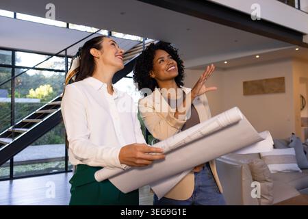 Two women in business-casual clothes holding rolled plans and pointing in living room near stairs Stock Photo