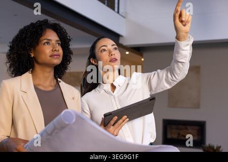 Diverse female coworkers holding blueprints and tablet, pointing and inspecting modern mezzanine Stock Photo