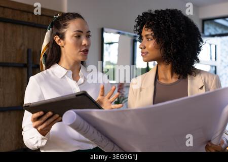 Diverse female colleagues in workwear discussing plans with tablet and rolled blueprints in office Stock Photo
