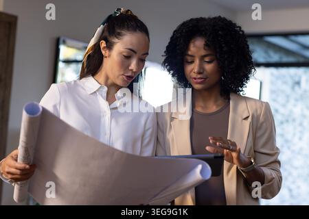 Diverse female coworkers in blazers reviewing large blueprints while using tablet in meeting room Stock Photo