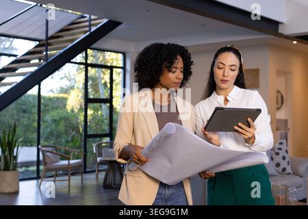 Diverse female colleagues reviewing rolled plan and tablet in business attire in modern home Stock Photo