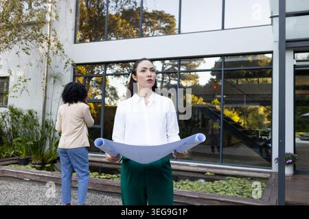 Diverse female coworkers standing in courtyard holding rolled plans by glass windows, white blouse Stock Photo