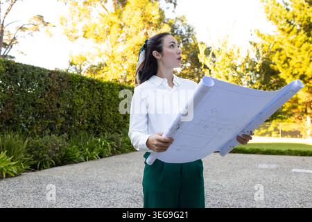 Woman inspecting rolled blueprints on driveway wearing white shirt green pants patterned scarf Stock Photo