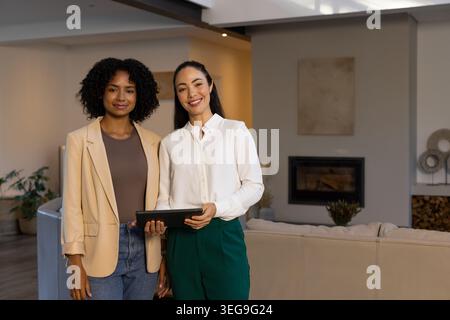 Diverse female coworkers standing in modern living room wearing business attire holding tablet Stock Photo