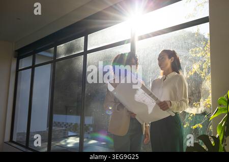 Diverse female coworkers in office wear examining plans near glass wall with lens flare, copy space Stock Photo