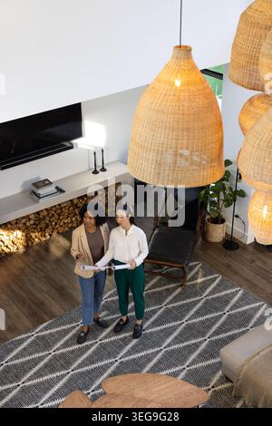 Diverse female coworkers reviewing rolled plans in modern living room with woven pendant lights Stock Photo