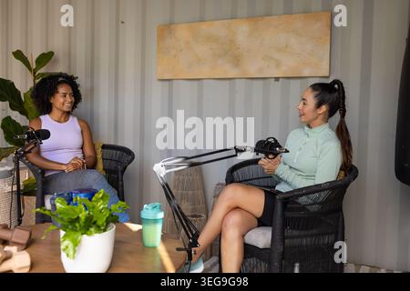 Diverse female cohosts sitting in chairs in small studio wearing tops recording podcast with mic Stock Photo