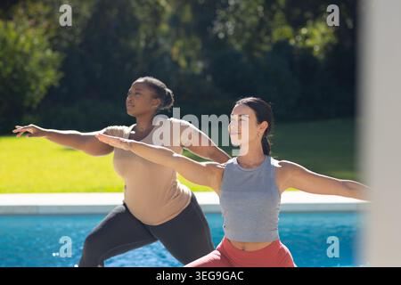 Diverse female friends practicing standing yoga in backyard beside swimming pool wearing leggings Stock Photo