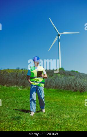 A windmill engineer stands in a green field with a wind turbine in the background. He is using a tablet to view data and monitor the turbine during da Stock Photo