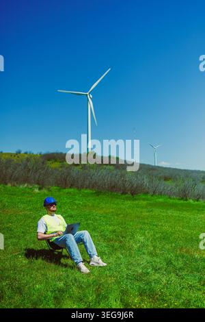 A windmill engineer sits on a chair in a grassy area using a laptop. Tall wind turbines stand in the background under a clear blue sky. The setting is Stock Photo