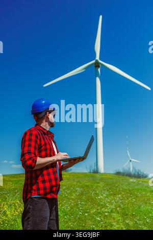 Engineer wearing a hard hat stands in field with laptop, focused on task related to wind turbine while enjoying clear sky and green grass around. Stock Photo