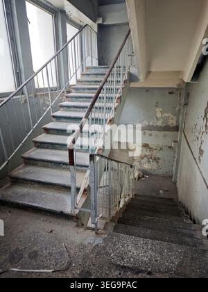 Dusty concrete stairwell in an abandoned building with peeling paint, worn railings and tall windows. Decayed interior architecture in natural light. Stock Photo