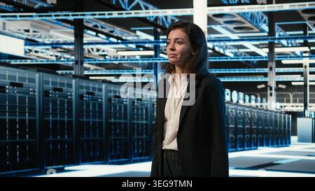Smiling data center manager taking virtual tour using AR technology, pleased by performance. Happy woman walking in server hub with blue wireframe mesh gear augmented reality simulation Stock Photo