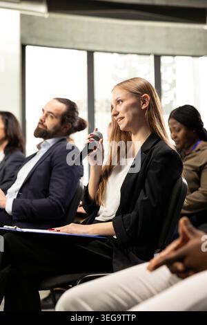 Young female journalist asking questions on microphone and taking notes, interacting with speaker and engaging in debate. International business summit with diverse participants in attendance. Stock Photo