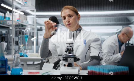 Female microscopist examining microscope glass slide in laboratory to view cells. Research facility employee checks sample slide, doing imaging process using optical device, camera A Stock Photo