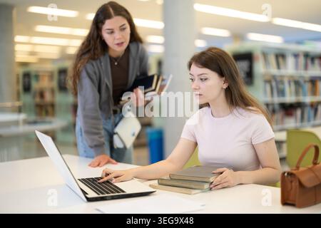Two girls studying for an exam together in university library using books and laptop Stock Photo