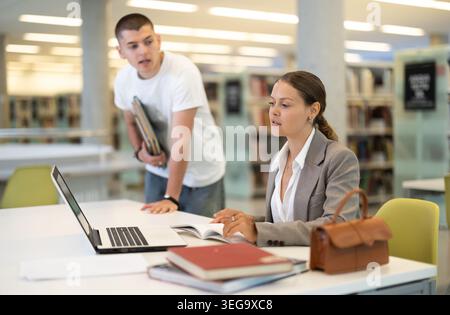 Girl and boy studying for exam together in library - using books and laptop Stock Photo