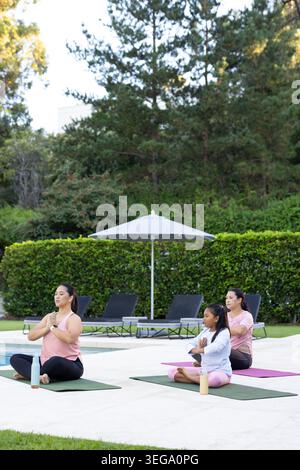 Diverse female group practicing seated yoga on poolside patio with colorful mats, bottles Stock Photo