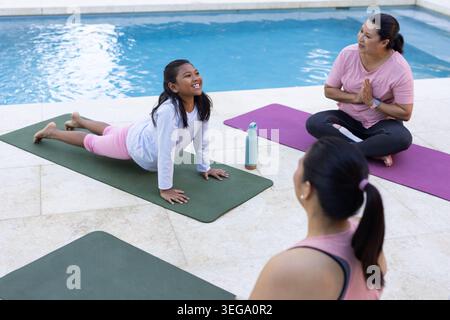 Diverse female coach and students doing yoga on pool deck with mats, water bottle, wearing leggings Stock Photo