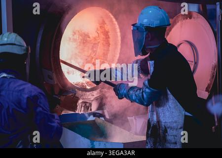 A worker in full protective gear, including a face shield and heat-resistant suit, uses a rod to probe inside a blazing hot foundry furnace. Stock Photo