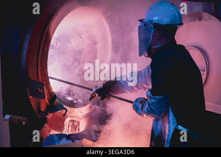 A worker in full protective gear, including a face shield and heat-resistant suit, uses a rod to probe inside a blazing hot foundry furnace. Stock Photo