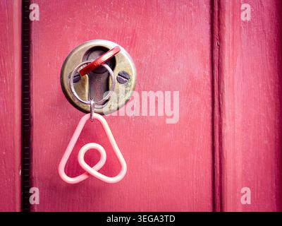 A close-up view of an Airbnb logo keychain hanging from a key inserted into a brass cylinder lock on a weathered pink wooden door. Stock Photo