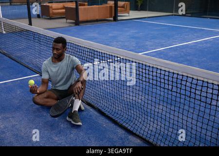 Sitting man wearing gray tee and black shorts holding yellow ball and paddle on blue court Stock Photo