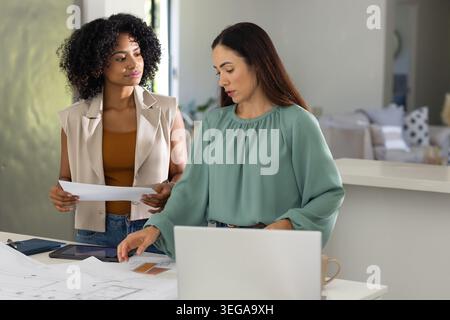 Diverse female coworkers standing at kitchen counter reviewing floor plans with laptop and swatches Stock Photo