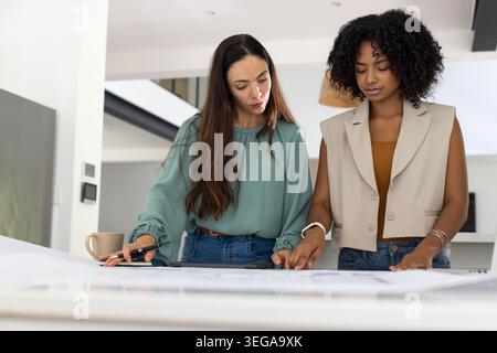 Two female colleagues leaning over large blueprints on table in bright studio, marking on tablet Stock Photo