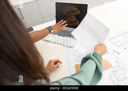 Diverse female coworkers reviewing blueprints on white table, pointing at laptop 3D wireframe Stock Photo