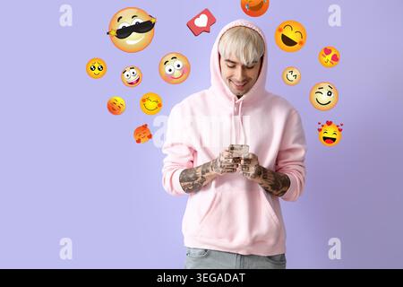 Young tattooed man in pink hoodie, with mobile phone and happy emojis on lilac background Stock Photo
