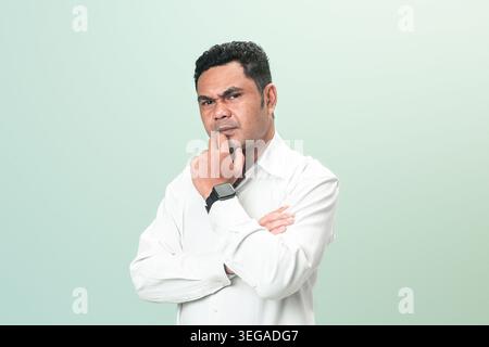 A serious Indonesian professional in a white shirt stands with crossed arms and touches his chin, thinking deeply against a green background. Stock Photo