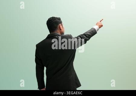 Back view of a professional man in a black suit pointing finger up and forward against a clean green background with blank space. Stock Photo