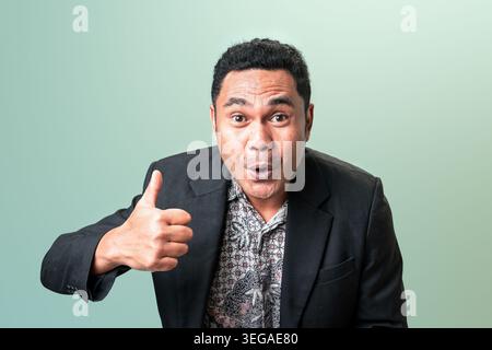 An amazed Indonesian man in a suit jacket and batik shirt gives a thumbs up with a surprised happy face against a green background with copy space. Stock Photo