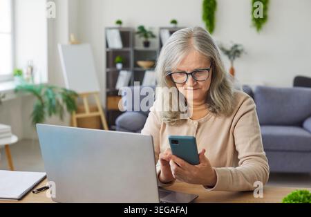 Old senior woman sitting at table holding smartphone looking down at screen, interacting online Stock Photo