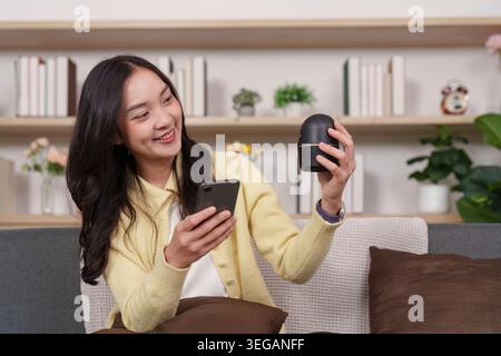 Smiling Asian woman holding smartphone and smart device in modern home Stock Photo