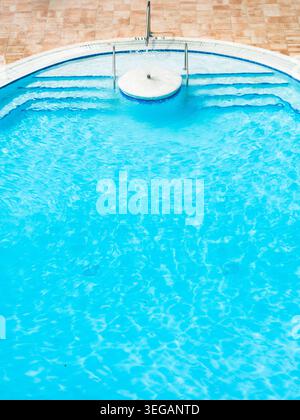 A recreational swimming pool features curved stairs and metal handrails, positioned next to a tan ceramic-tiled floor. Stock Photo