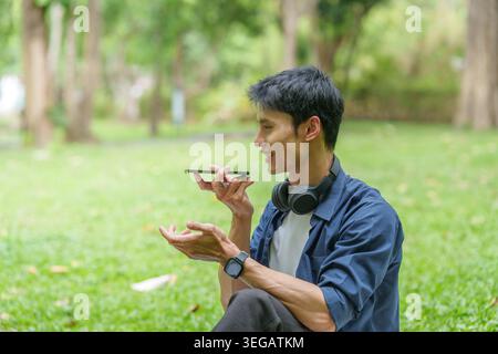 Asian man using AI voice assistant on smartphone in outdoor park Stock Photo
