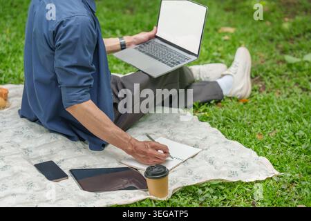 Asian man working remotely on laptop and writing in notebook in outdoor park Stock Photo