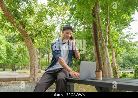 Asian man working remotely on laptop and talking on smartphone in outdoor park Stock Photo
