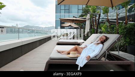 Woman in white bathrobe relaxing and smiling on a sun lounger at a luxurious rooftop terrace overlooking Hong Kong's vibrant Victoria Harbour skyline. Stock Photo
