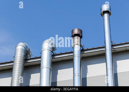 Shiny stainless steel ventilation chimneys and exhaust pipes rise from an industrial rooftop; reflecting sunlight against a blue sky. Modern exterior Stock Photo