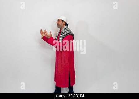 Indian man wearing traditional Muslim clothing, including a red long tunic and a white cap with a checkered scarf. He is holding prayer beads Stock Photo