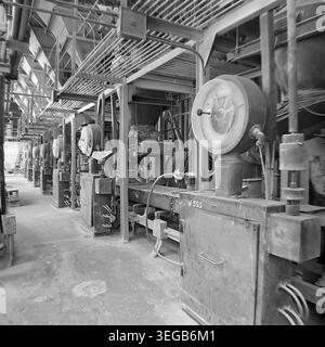 Sloviansk, 1981, AIZ (Armature-Insulator Plant). A perspective view down a row of massive industrial hoppers and mixing mechanisms inside the factory. Large circular dials and gauges are attached to the heavy metal equipment, highlighting the technical monitoring of the manufacturing process. A historical black and white archive photograph capturing the authentic atmosphere of the era, the complex engineering infrastructure, and the plant interior. A detailed glimpse into the past, Soviet industry, and the peaceful Donbas. Stock Photo