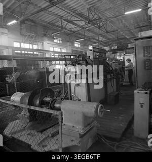 Sloviansk, 1981, AIZ (Armature-Insulator Plant). A male worker stands at the control area of a heavy industrial machine inside a bright factory workshop. In the foreground, a large mechanical spool setup is partially enclosed by a protective wire mesh fence, indicating a specialized manufacturing stage. A historical black and white archive photograph capturing the authentic atmosphere of the era, the engineering infrastructure, and everyday life. A detailed glimpse into the past, Soviet industry, and the peaceful Donbas. Stock Photo