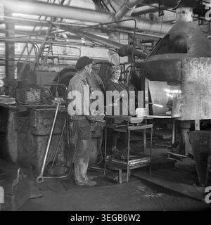 Sloviansk, 1981, AIZ (Armature-Insulator Plant). A male worker in a flat cap and thick protective gloves stands alongside a female colleague near a brightly glowing industrial furnace. They are carefully using long metal tools to handle materials inside the intensely hot thermal processing unit within the busy manufacturing workshop. A historical black and white archive photograph capturing the authentic atmosphere of the era, the hazardous metallurgical working conditions, and the peaceful Donbas. Stock Photo